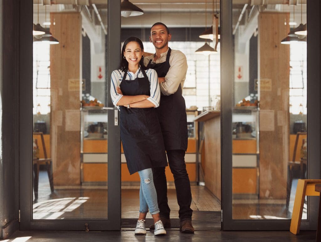 Proud business partners standing in their supermarket entrance. Business colleagues standing in their coffeeshop. Portrait of married couple working in cafe together. Colleagues arms crossed in shop